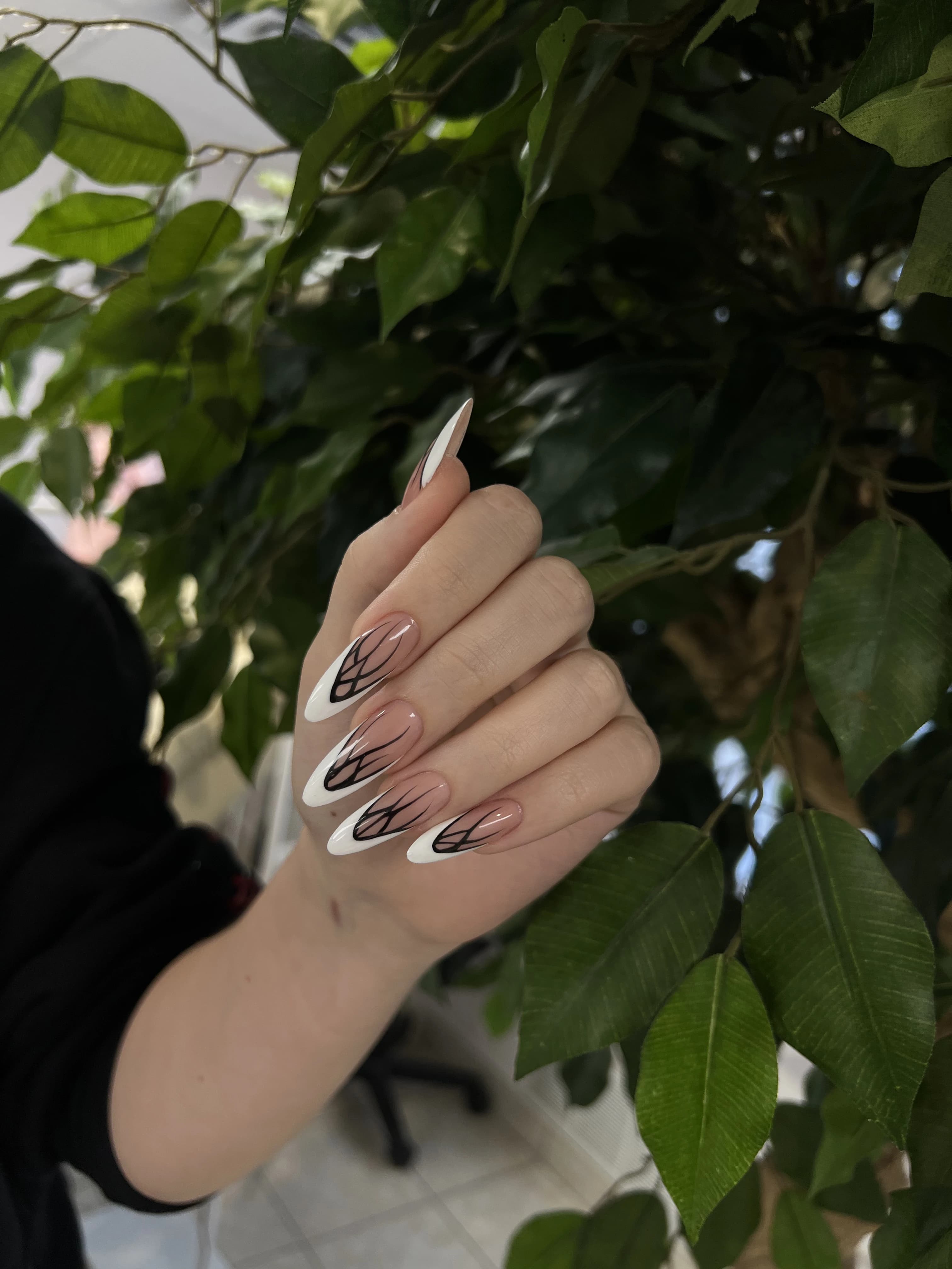 Gel-X extensions at My Beauty Space: long almond French tips with black curved line art, posed before green foliage.